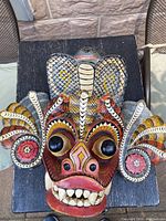 Front view of a colorful wooden mask featuring a red face with large eyes and white teeth, decorated with snakes on the top and sides.