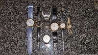 Top-down view of seven wristwatches placed on carpet background. Shows different strap materials and face designs for identification.