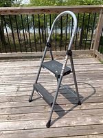 Front and side views of a two-step folding stool with a rounded handle, metal frame and textured steps