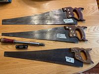 Photo showing three 26-inch crosscut hand saws with wooden handles and labels indicating brand and teeth per inch parameters along with ratcheting screwdriver.
