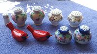 Eight assorted salt and pepper shakers arranged on a blue surface, showing floral patterns, bird shapes, and varying ceramic designs.