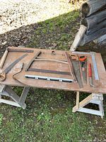Photo showing various woodworking hand tools on a wooden board including saws, levels, and a nail puller outdoors on grass