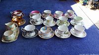 Overview of 12 teacups and 12 saucers in various floral and decorative patterns with a jug and tray on blue carpet under natural light.
