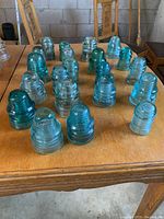 Front overview of 23 glass insulators arranged on wooden table showing different shapes and colors in blue/aqua hues.