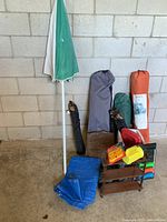 Camping equipment including tents in carry bags, sun umbrella, portable chairs in bags, tarp ground sheet, and cooler with bbq lighters.