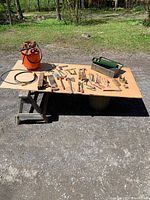 Overview photo showing all items on plywood table outdoors, including orange air pig, toolbox, and various hand tools.
