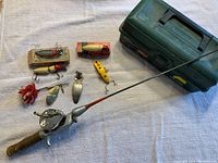 Photo showing the full fishing rod, Ocean City 1600 reel mounted, green plastic tackle box, and seven fishing lures arranged on a light colored cloth.