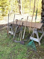 Photo showing eight yard tools including rakes, pitchforks, a hoe, a scythe, and pruning shears arranged leaning against a wooden workbench outdoors.