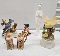 Photo showing a collection of bird figurines, two stein salt and pepper shakers, a colorful bird music box, and a white praying statue against a plain background.