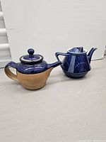 Two ceramic teapots on a gray surface against a plain background, showing detailed blue decorations with one natural beige base and the other fully decorated in blue patterns.