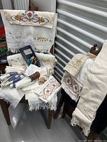 Multiple vintage linens arranged on chairs showing embroidery, lace edging, fringed hems, and folded pieces.