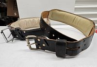 Two black leather weight lifting belts with silver metal buckles laid side by side showing padded interior.
