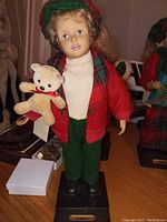 Musical Christmas boy figure approximately 16 inches tall, wearing green pants, red jacket, white turtleneck and plaid hat, holding teddy bear, mounted on a black NOEL base. Shows signs of smoke damage, untested.