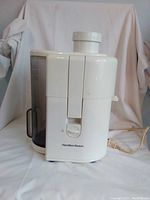 Side view of white Hamilton Beach electric juicer showing juice collection container and power cord, placed against a white backdrop.