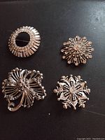 Photo showing four vintage silver-tone brooches with clear rhinestones, various floral and abstract designs on a dark background.