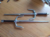 Two metal sai weapons laid flat on a wooden floor, showing the polished octagonal shafts and curved prongs. One sai has a leather wrapped handle grip, the other handle is bare metal.