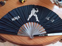 Black folding fan featuring white ninja illustrations and text placed on a round wooden table.
