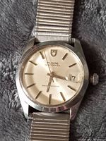 Front view of the watch showing silver dial, baton markers, date at 3 o’clock, acrylic crystal scratches and expansion band