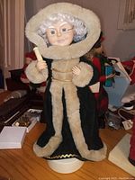Photo of a vintage Mrs. Santa Claus figurine standing on a white round base. The figurine wears a green velvet coat with beige faux fur trim and holds a scroll in one hand. Facial features are molded with curly white hair. Missing glasses as noted.