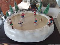 Top angle view of vintage skating pond showing five ice skating figurines on ice surface with surrounding trees and lamppost, visible wear and discoloration on plastic surface.