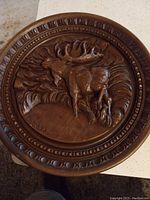 Photo shows round wooden plaque featuring a carved moose with detailed antlers and forest background, surrounded by a decorative circular border.