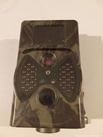 Front view of the hunting trail camera in a camouflage case showing lens, infrared LEDs, and display screen.