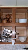 Wooden kitchen cabinet shelf containing a collection of woven bread baskets, ceramic bowls, and various metal cake pans and muffin tins stacked haphazardly.
