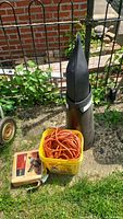 Sears electric drill box, orange extension cord coiled inside yellow bucket, and tall dark convertible boot storage container on grass by garden fence.