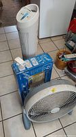 Photo showing all three fans: white standing tower fan, boxed window fan, and round floor fan on the tiled floor.