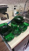 Multiple pieces of forest green glass bowls and plates displayed on kitchen counter next to sink with soap and faucet in background.