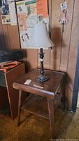 Wooden side table with dark finish and visible scratches supports a tall metal table lamp with beige fabric shade.