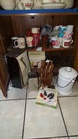 Shelf with assorted ceramic coffee mugs, to the right a white compost jar with lid and handle, in front grater, knife block with knives and other utensils