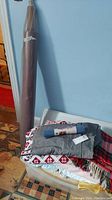 Photo showing bolt of brown fabric leaning against the wall next to a plastic container of folded blankets and linens including a gray blanket with red and white patterns and a red and black plaid blanket.