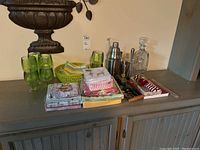 Photo showing an overview of the lot on a wooden cabinet including green glassware, cocktail shaker, clear glass decanter, bottle openers, lemon squeezer, napkins and towels in basket.