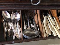 Open drawer showing silver plate and stainless steel spoons, ladles, and knives with white handles organized in compartments.