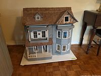 Front view of the large handmade wooden dollhouse painted blue with white trim and brown shingled roof. Shows architectural details including windows with shutters and wreaths, and porch with columns.