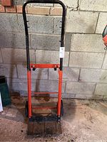 Front view of metal dolly leaning against a gray concrete wall, showing the handle and base platform.