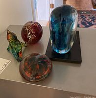 Photo showing four glass items including green marine animal figure, red and white orb, clear glass orb with red and black design, and blue glass award on black base.