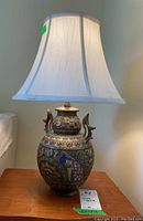 Overall view of the table lamp showing the white fabric shade and painted metal base with colorful, intricate designs and sculptural handles.