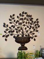 Front view of metal foliage planter wall art mounted on a white wall above kitchen or dining area with green glassware below.