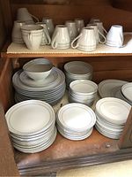 Full view of various stacked Mikasa Nordica china pieces including cups, plates, and bowls inside wooden cabinet shelf