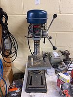 Front view of the Ryobi DP100 drill press showing the metal base, worktable, and blue motor cover with control label.