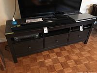 Front view of dark brown TV/media stand with three drawers and two open shelves, with television and audio equipment on top.