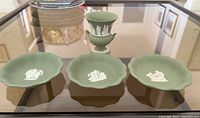 Photo showing three green Wedgwood Jasperware trinket plates and one urn, displayed on glass table, highlighting white classical relief details on each item.