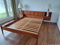 Full view of teak bed frame showing slatted support, headboard with attached side drawers and two lamps