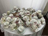 Group photo showing more than 35 single tea cups and matching saucers arranged on a table. Various floral and patterned designs, mostly white with colorful flowers and gold trim.