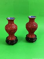 Pair of cinnabar lacquer vases with blue enamel interior on black wooden stands against green background.
