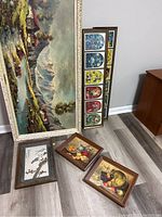 Photo showing large framed mountain landscape canvas leaning against wall, multipane frame with stained glass window prints, and three smaller framed still life paintings on floor.