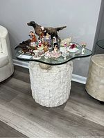 Full view of the artistic side table showing the rough white stone veneer cylindrical base topped with an irregular shaped clear glass top holding several small ceramic figurines and collectibles.