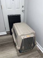 Plastic dog crate with metal wire door and foldable wire dog crate behind it, placed near a white door.